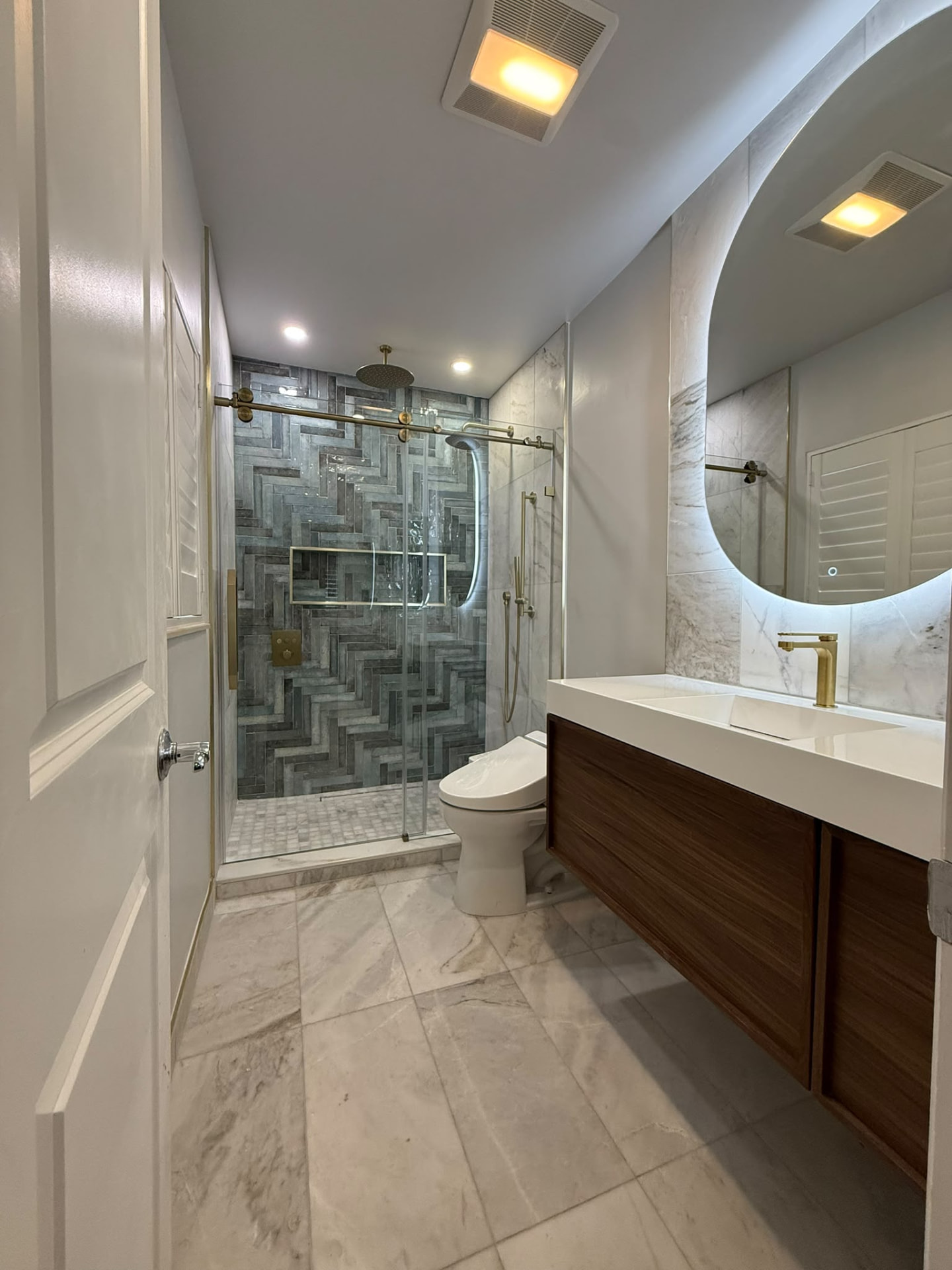 Completed bathroom with a gray herringbone tile shower wall, brushed-gold sliding glass door hardware, backlit round mirror, and a walnut floating vanity